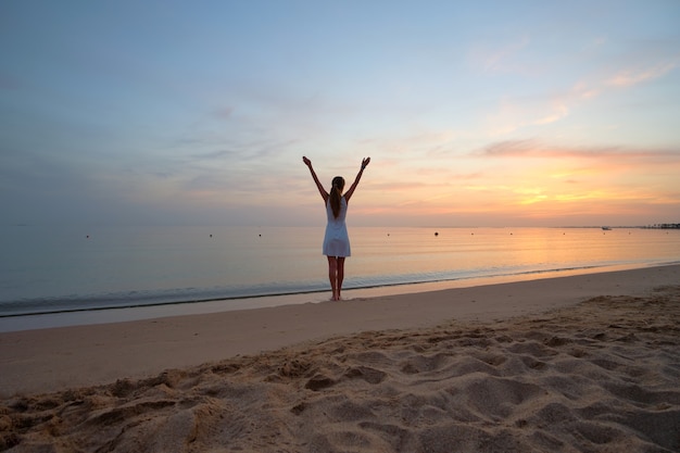 Young happy woman standing on sandy beach by seaside enjoying warm tropical evening.