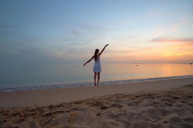 Young happy woman standing on sandy beach by seaside enjoying warm tropical evening.