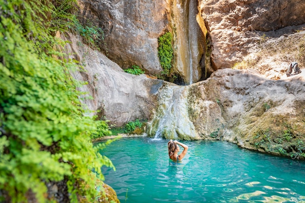 Молодая счастливая женщина плавает в бирюзово-голубой кристально чистой воде в реке с водопадом