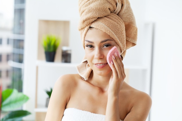 Young happy woman in towel making facial massage with organic face scrub in stylish bathroom