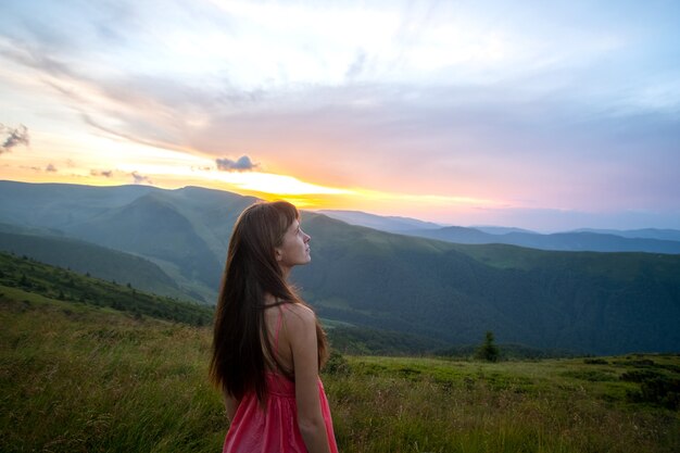 日没時の自然の景色を楽しむ夏の山々の風の強い夜に草が茂った丘の中腹に立っている赤いドレスを着た若い幸せな女性旅行者。