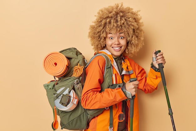 Young happy woman uses trekking poles for walking in mountains carries rucksack with necessary equipment dressed in orange windbreaker isolated over brown background Tourist traveler indoor