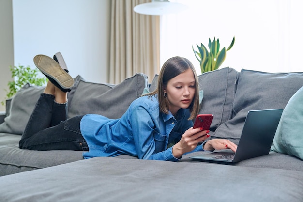 Young happy woman using smartphone laptop lying on sofa at home