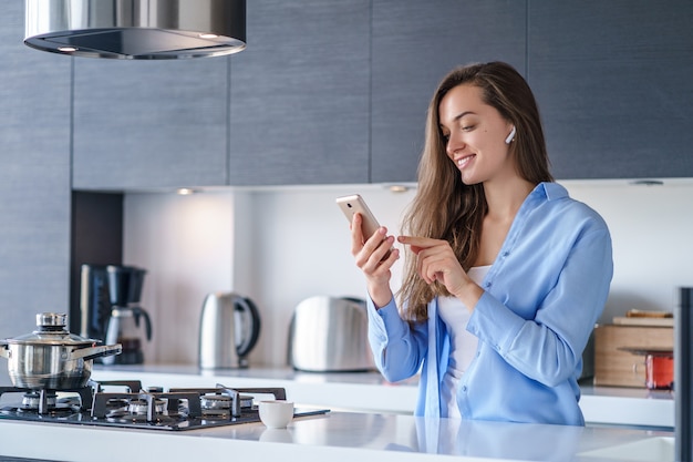 Young happy woman using smartphone and wireless headphones for listening music and making video calls in the kitchen at home. Modern mobile people