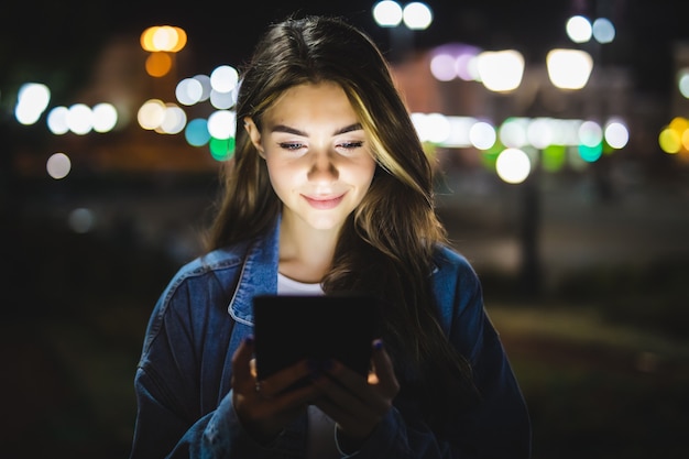 Young happy woman using tablet outdoors over blurry night city lights