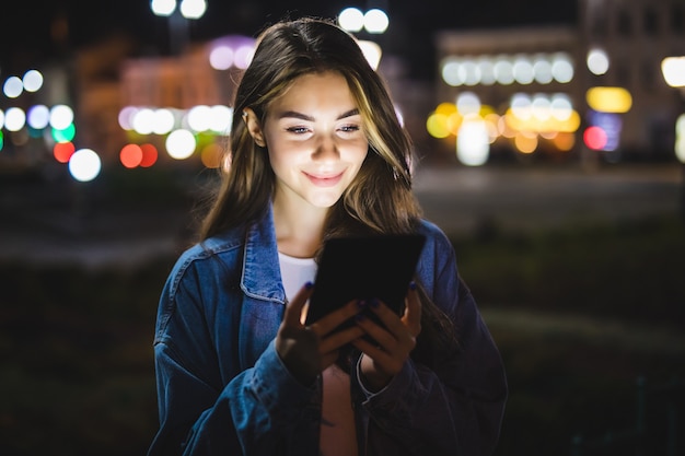 ぼやけた夜の街の明かりの上で屋外でタブレットを使用して若い幸せな女性