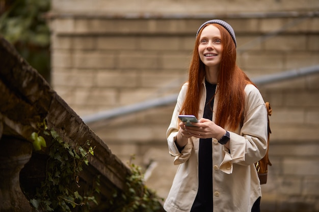 Young happy woman walking in historical place using smartphone, using mobile phone, portrait of redhead lady in coat alone outdoors, going to take photo