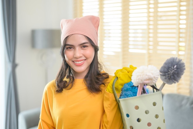 Young happy woman wearing yellow gloves and holding a basket of cleaning supplies in living room