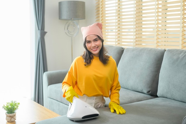Молодая счастливая женщина в желтых перчатках и пылесосом чистит диван в гостиной