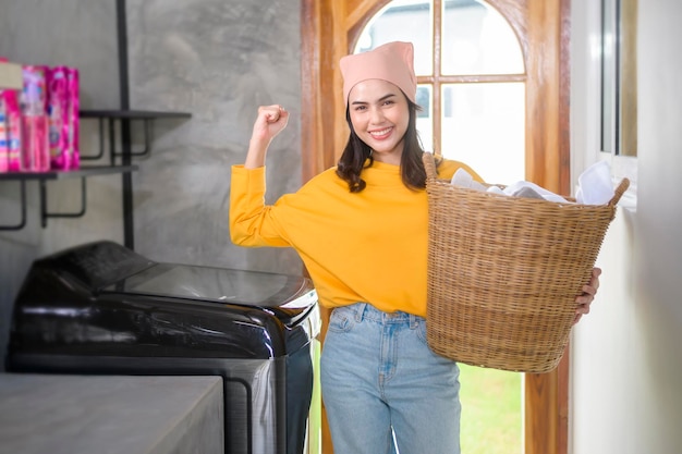 Молодая счастливая женщина в желтой рубашке держит корзину с одеждой в домашней прачечной