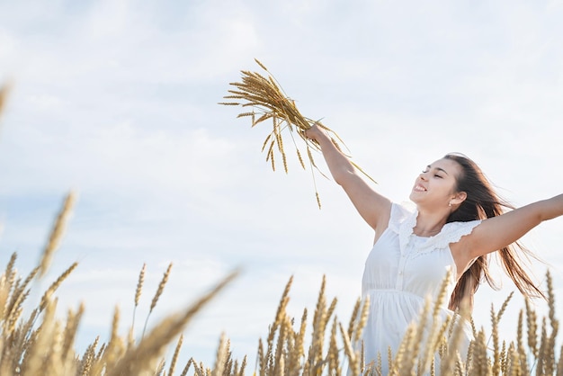 Young happy woman in white dress standing on a wheat field with\
sunrise on the background