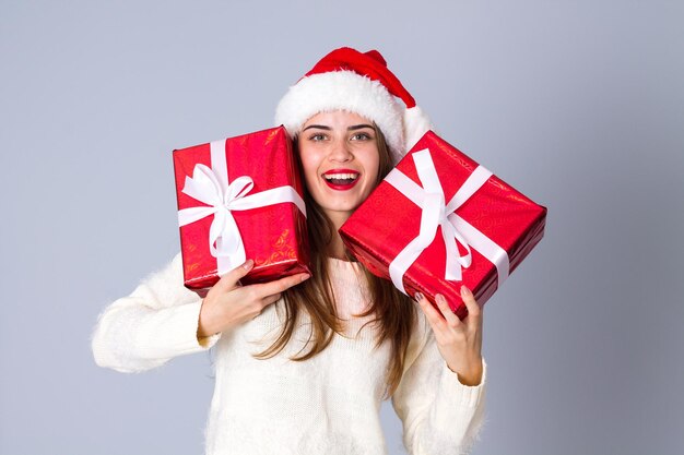 Young happy woman in white sweater with red christmas hat holding presents on grey background