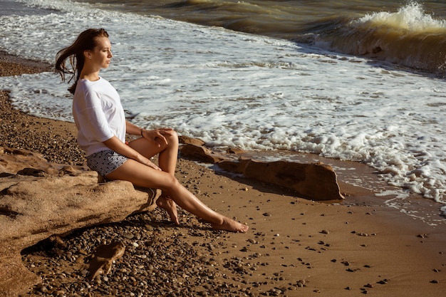 Young happy woman in white t-shirt and shorts sits on the seashore in summer