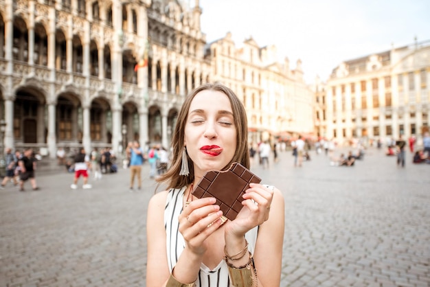 ベルギーのブリュッセルのグランドプレイスに屋外に立っているダークチョコレートバーを持つ若くて幸せな女性。ベルギーはチョコレートで有名です