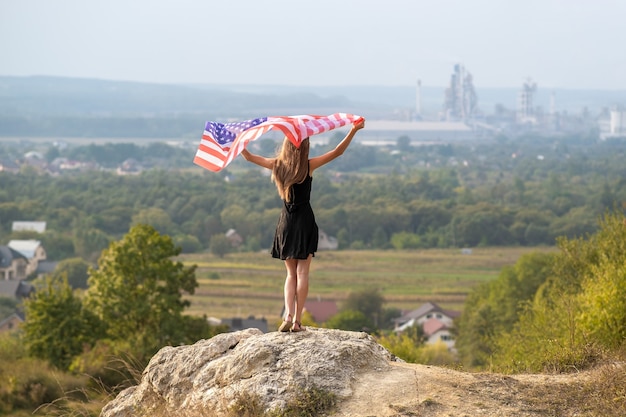 Foto giovane donna felice con i capelli lunghi che si alzano sventolando la bandiera nazionale americana del vento nelle sue mani in piedi su un'alta collina rocciosa godendosi la calda giornata estiva.