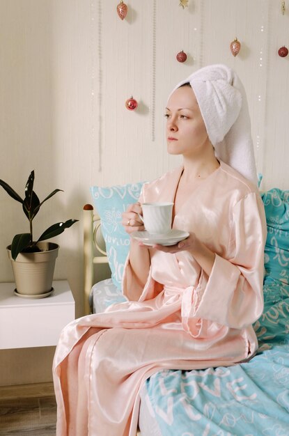 Young happy woman with a towel on her head sitting in bed in pajamas in a cozy home. Drinking coffee