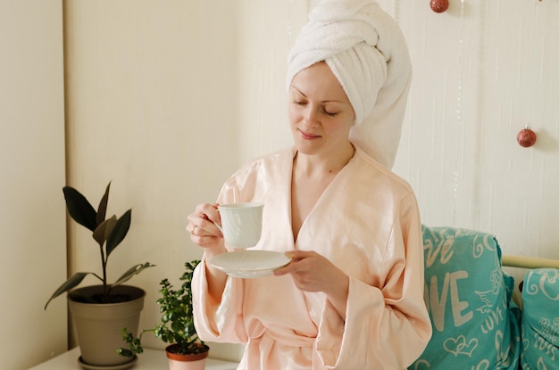 Young happy woman with a towel on her head sitting in bed in pajamas in a cozy home. Drinking coffee