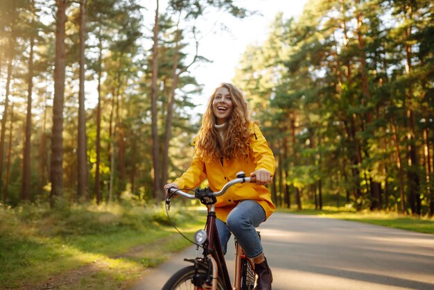 노란색 코트를 입은 젊은 행복한 여성은 햇볕이 잘 드는 공원에서 자전거를 타고 휴식을 취합니다.
