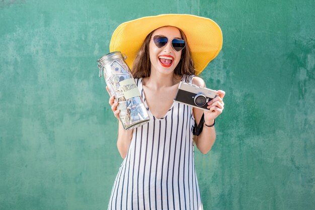 Молодая и счастливая женщина в желтой шляпе держит банку, полную денег для путешествия на зеленом фоне. Экономия денег для концепции летних каникул