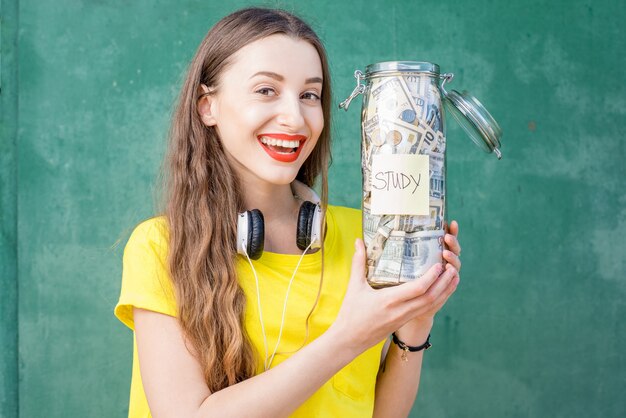 Молодая и счастливая женщина в желтой футболке держит бутылку с деньгами для учебы