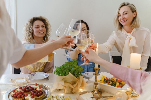 Amici di giovani donne felici che tintinnano i bicchieri e sorridono al tavolo festivo servito con cibo e vino bianco decorato con candele