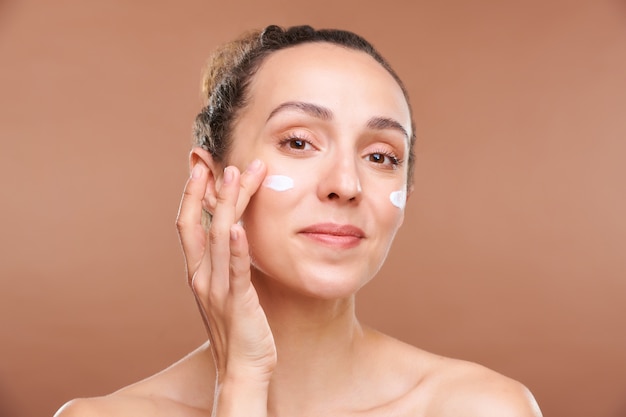 Young healthy brunette female looking at you while taking care of face and applying rejuvenating cream after removing makeup