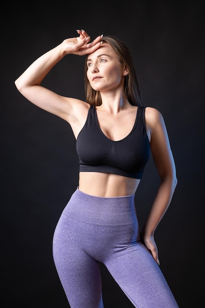 A young healthy girl in a sports top and leggings poses in the studio against a black background