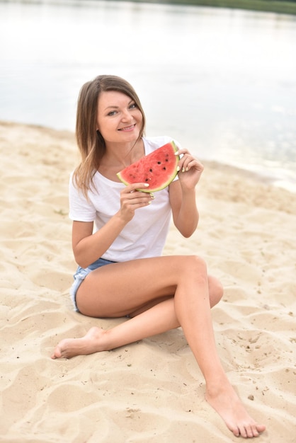 白いTシャツの若い健康なスリムな女性はスイカのスライスを保持し、ビーチで夏を楽しんでいます