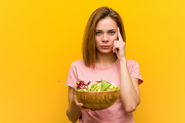 Фото Молодая здоровая женщина держа салат указывая ее висок с пальцем, думая, сфокусированный на задаче.
