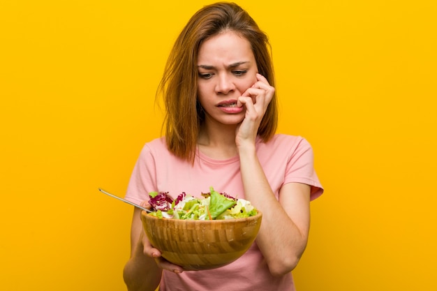 Young healthy woman holding a salad biting fingernails, nervous and very anxious.