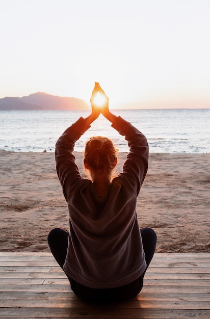 Young healthy woman practicing yoga holding hands in meditation\
pose with sun between themon the bea...