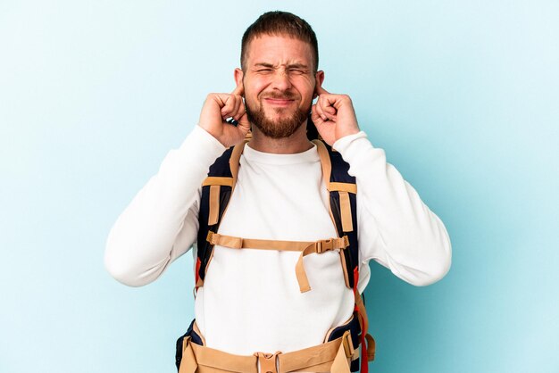 Photo young hiker caucasian man isolated on blue background covering ears with hands.