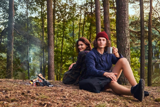 Young hiker couple sitting back to back while warming near a\
campfire at camp in the forest. travel, tourism, and hike\
concept.