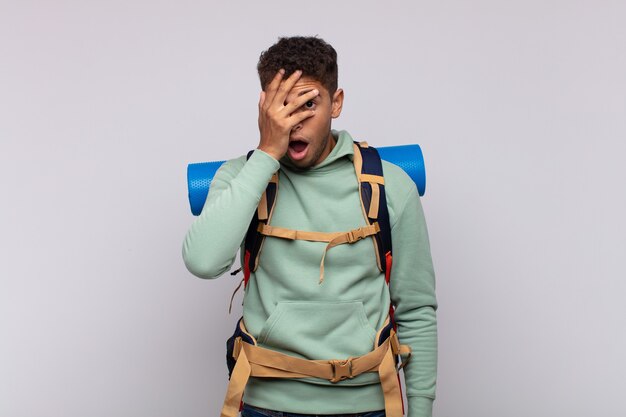 Young hiker man looking shocked, scared or terrified, covering face with hand and peeking between fingers