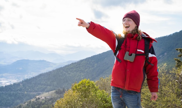 Young hiker man with backpack on top of mountain pointing with hand to horizon and smiling