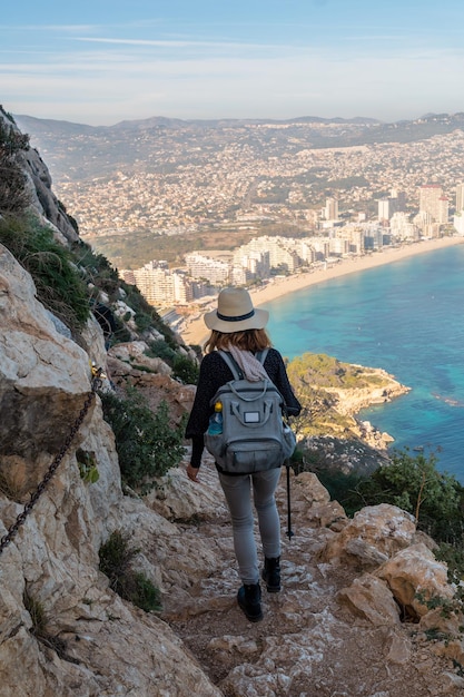 A young hiker wearing a hat on the descent path of the Penon de Ifach Natural Park with the city of Calpe in the background Valencia Spain Mediterranean sea View of La Fossa beach