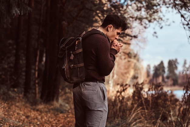 A young hiker wearing sweater warms hands in the cold autumn forest on a cloudy day.