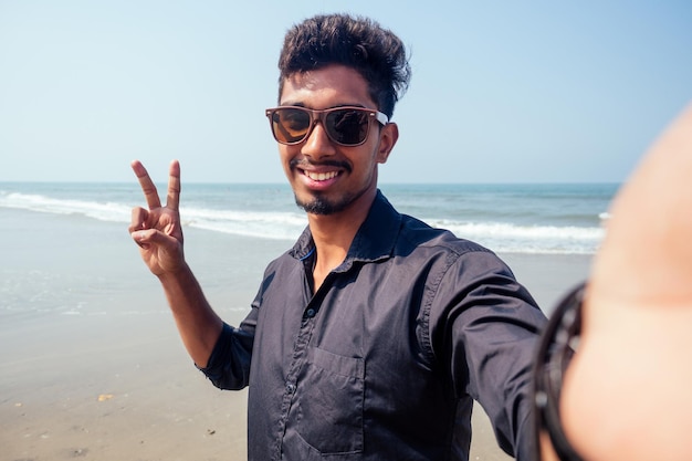 Giovane uomo alla moda indù che scatta una foto autoritratto sulla fotocamera frontale dello smartphone con occhiali da sole vacanza al mare attiva sulla spiaggia felice di goa india. concetto di protezione solare spf.