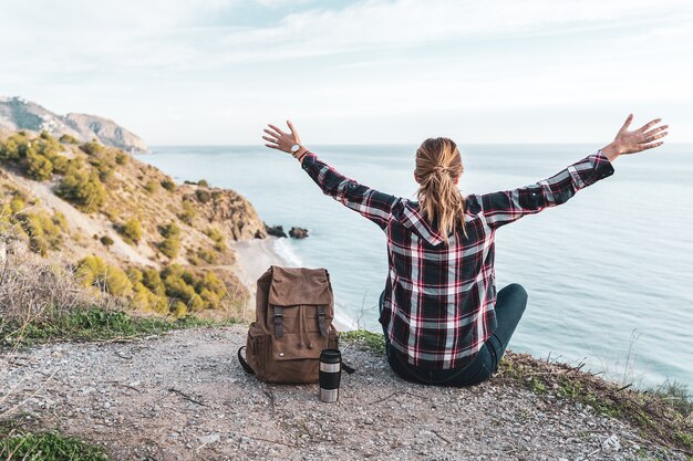 Young hip woman with open arms and a backpack explores the coast on a beautiful day. Concept of exploration and adventures