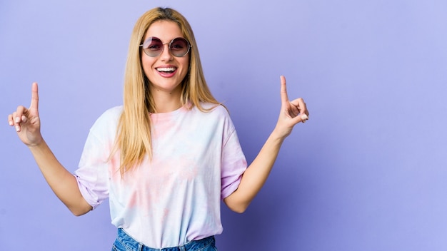 Young hipie woman with glasses isolated on purple background indicates with both fore fingers up showing a blank space.