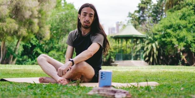 Молодой хиппи-латиноамериканец разговаривает по видеосвязи, сидя в позе лотоса на коврике для йоги на траве с беседкой позади