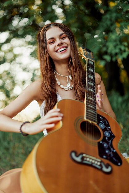 A young hippie woman with a guitar in her hands smiles sweetly into the camera on a trip to nature lifestyle in harmony