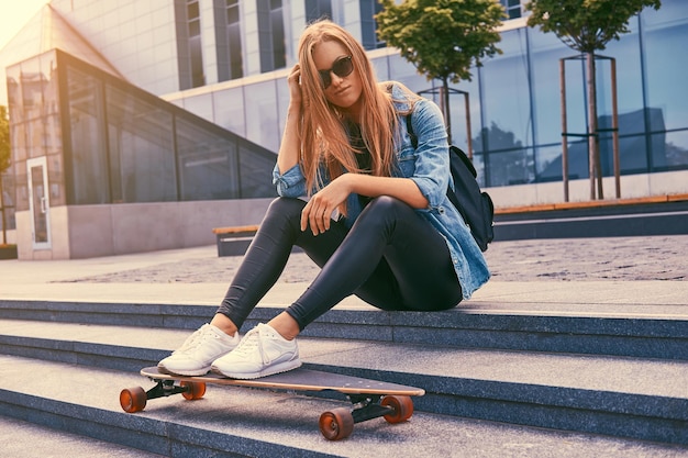 Ragazza bionda giovane hipster in abiti casual e occhiali da sole, seduta sui gradini contro un grattacielo, riposando dopo aver cavalcato su uno skateboard.