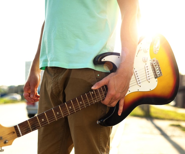 Молодой хипстер в синей футболке с гитарой стоит на дороге в отличие от солнечного света и бликов
