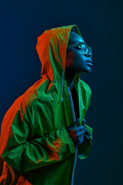 暗いスタジオの背景側のビューの肖像画の上にネオン照明に立っているレインコートを着ている若い流行に敏感なファッション女性