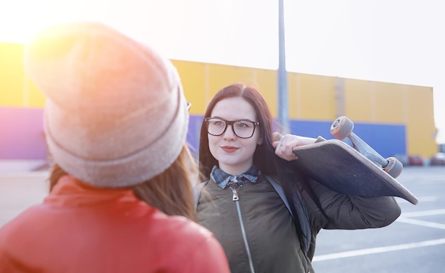 流行に敏感な少女がスケートボードに乗っています。スケートボードを持って街を散歩する女の子のガールフレンド。スケートボードで路上で春のスポーツ。