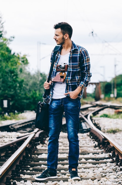 Young hipster guy tourist with camera and bag standing on the railroad