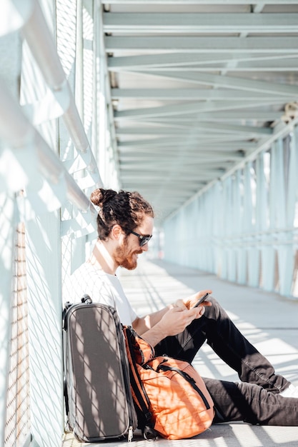 Young hipster man at the airport or bus station waiting while\
writes a text message with the luggage, bags and suitcase. young\
man long hair hipster traveler with sunglasses, copy space, sunny\
day