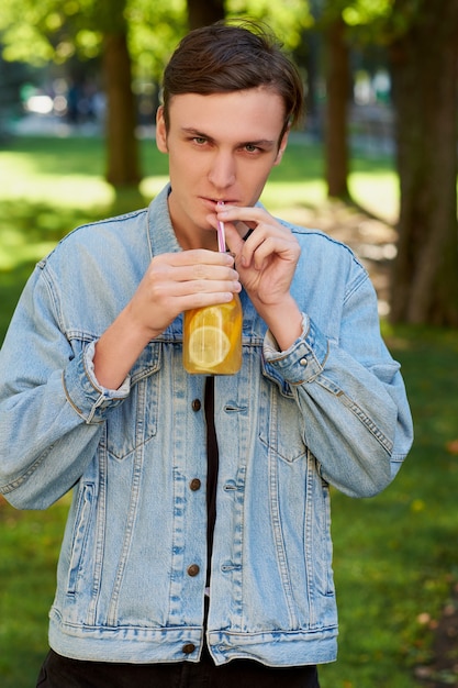 Young hipster man drinks healthy detox tea with citrus juice on green park background.