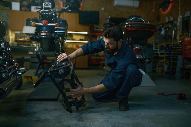 Young hipster man mechanic repairing motorcycle in workshop working with frame. Repairman checking and diagnosing parts on motorbike